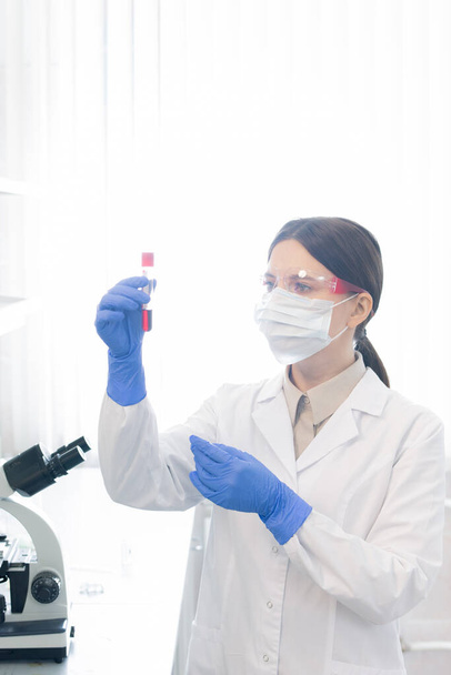 Vertical ,edium portrait of unrecognizable female pharmacologist wearing lab coat, protective mask. gloves and eyewear holding test tube with red liquid - 写真・画像