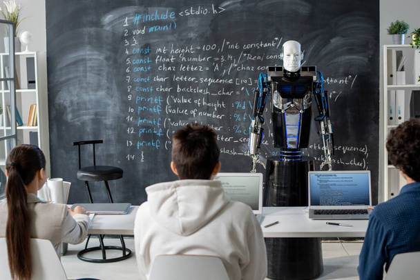 Zadní pohled na řadu studentů sedících u psacího stolu před automatizačním robotem stojícím u tabule místo lidského učitele - Fotografie, Obrázek