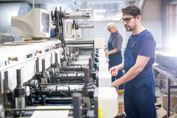 Serious young bearded employee of printing plant in blue overall working with modern machine - Foto, afbeelding