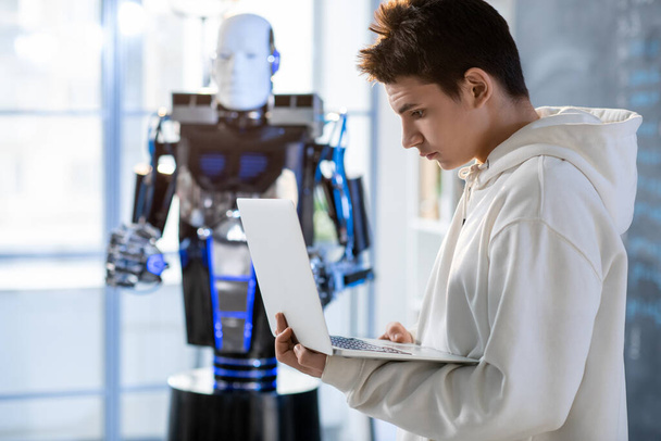 Νεαρός φοιτητής που εξετάζει την επίδειξη φορητών υπολογιστών ενώ εργάζεται με ρομπότ αυτοματισμού στο εργαστήριο ή στην τάξη του σύγχρονου ινστιτούτου - Φωτογραφία, εικόνα