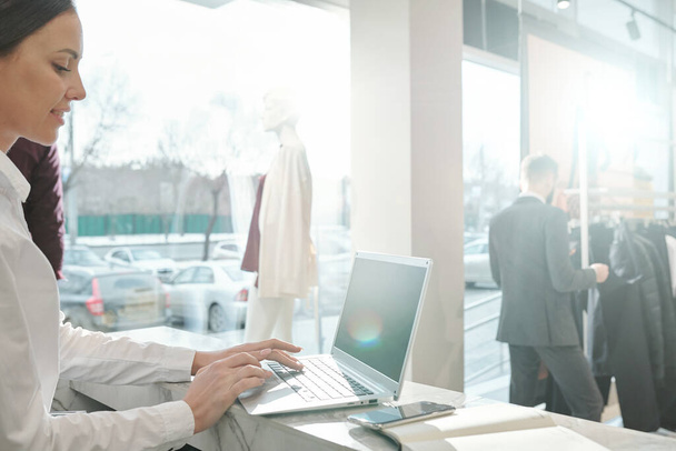 Hymyilevä nuori myyntipäällikkö seisoo tiskillä ja käyttää kannettavaa tietokonetta käsitellessään online-hakemuksia vaatekaupassa
 - Valokuva, kuva