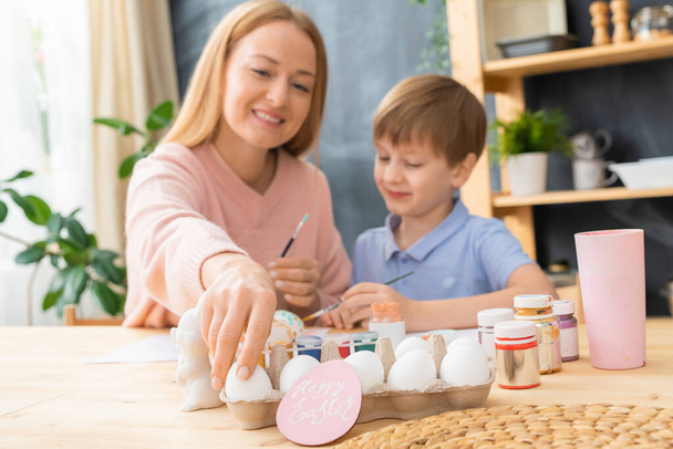 Smiling young mother and her son sitting at table and enjoying to paint Easter eggs - Foto, Bild