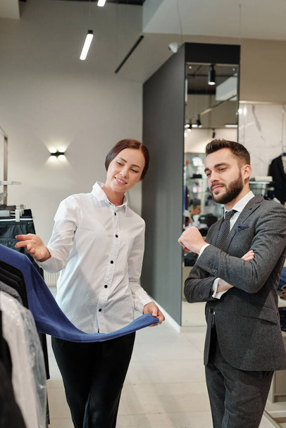 Φιλικός σύμβουλος καταστημάτων ενδυμάτων σε μπλούζα επιδεικνύοντας τη νέα συλλογή ρούχων ενώ συνεργάζεται με τον πελάτη στο κατάστημα - Φωτογραφία, εικόνα