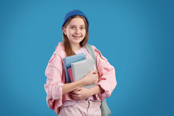 Портрет улыбающейся привлекательной девушки с брекетами, стоящими на синем фоне и держащими кучу учебников
 - Фото, изображение