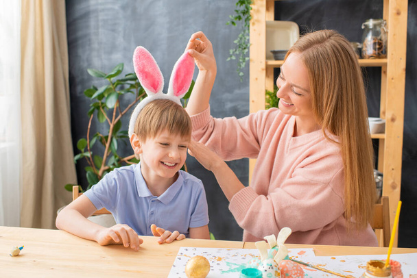 Szczęśliwa młoda matka dostosowując uszy królika na głowie syna podczas ubierania się na Wielkanoc - Zdjęcie, obraz