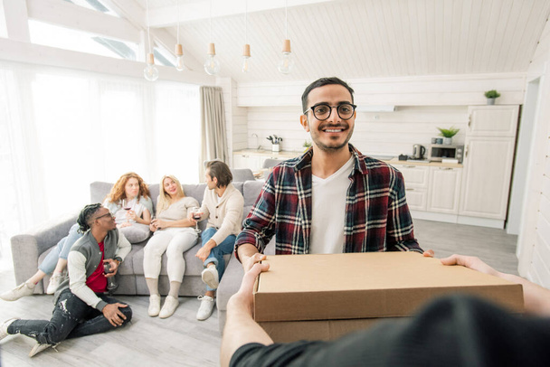 Молодому ближневосточному мужчине доставляют пиццу для друзей, собирающихся в современной квартире, горизонтальный снимок
 - Фото, изображение