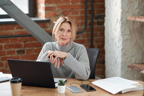 Портрет содержательной дамы-босса в сером свитере, сидящей за столом с ноутбуком в современном лофт-офисе
 - Фото, изображение