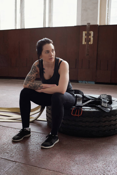 Νεαρή Καυκάσια αθλήτρια με τατουάζ που κάθεται πάνω σε λάστιχο και ξεκουράζεται μετά από προπόνηση στο γυμναστήριο - Φωτογραφία, εικόνα