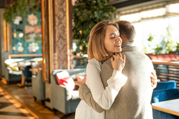 Jovem feliz e mulher apaixonados cumprimentando uns aos outros com abraço no restaurante moderno, tiro médio horizontal
 - Foto, Imagem