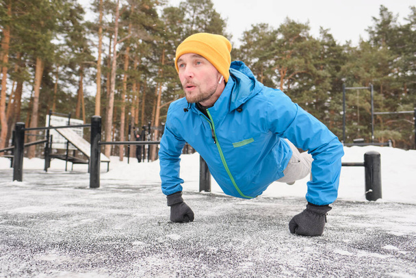 Kulaklık takan güçlü genç adam kışın antrenman alanında antrenman yaparken kendini yerden uzaklaştırıyor. - Fotoğraf, Görsel