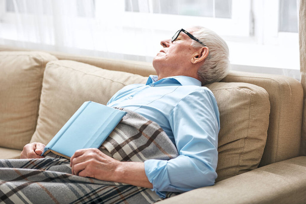 Fáradt idős férfi borított kockás szundikálás kanapén nyitott könyv, hogy nem fejezte be, és elaludt, miközben otthon maradt - Fotó, kép
