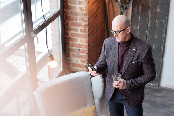 Acima vista de homem de meia-idade grave na jaqueta da moda usando gadget e beber álcool
 - Foto, Imagem