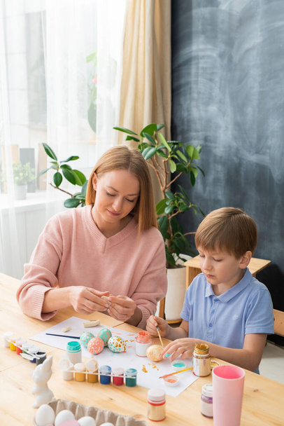 Zawartość młoda matka w różowym swetrze siedzi przy drewnianym stole z narzędziami artystycznymi i robi dekoracje wielkanocne z synem - Zdjęcie, obraz