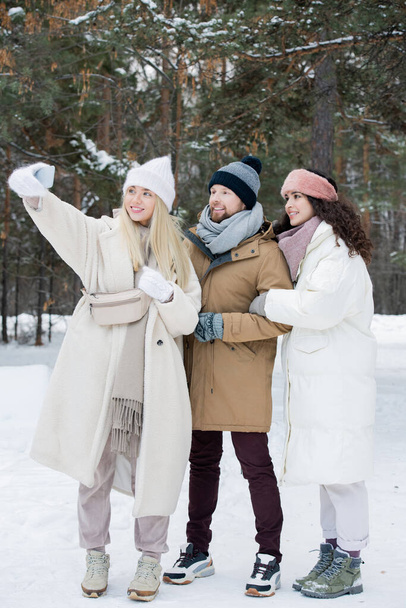 Pystysuora laukaus nuori mies ja kaksi naista viettää aikaa yhdessä ulkona talvipäivänä ottaen selfie kuva älypuhelimella
 - Valokuva, kuva