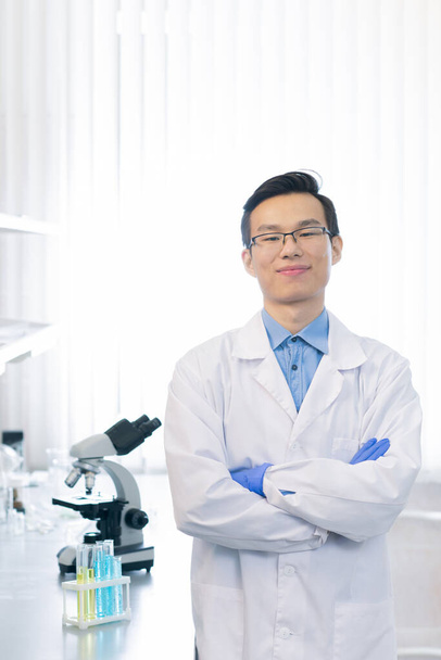 Fiducioso farmacologo asiatico che indossa camice da laboratorio bianco e occhiali in piedi con le braccia incrociate guardando la fotocamera sorridente in laboratorio moderno
 - Foto, immagini