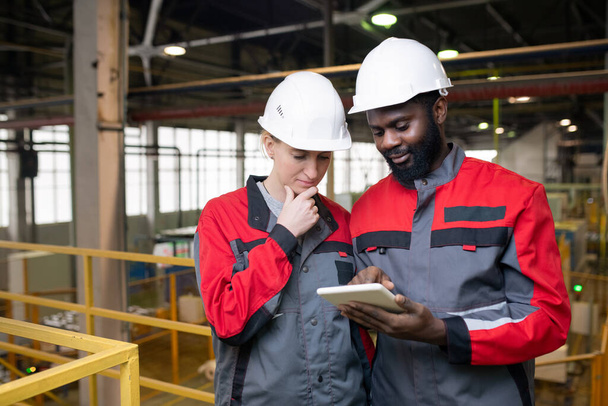 Jovens trabalhadores multiétnicos cuidadosos em capacetes usando tablet ao analisar dados de produção industrial
 - Foto, Imagem