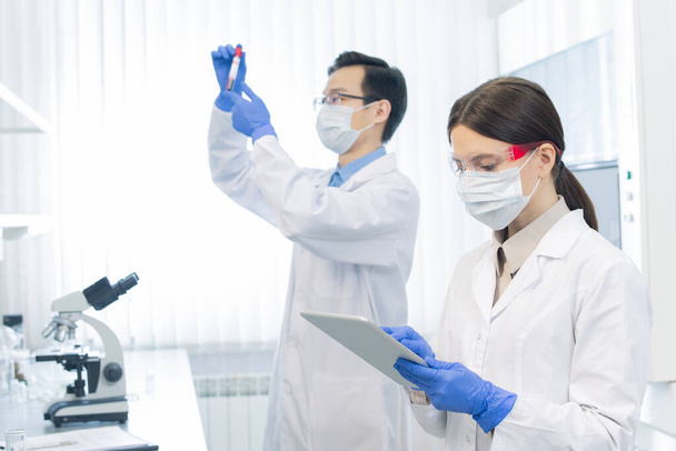 Два молодых учёных в защитных масках и перчатках работают над новыми лекарствами в современной лаборатории
 - Фото, изображение