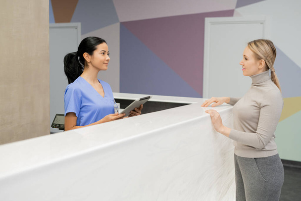 Mladá asistentka v modré uniformě stojící u přepážky recepce a konzultující pacientku v salonku stomatologických klinik - Fotografie, Obrázek