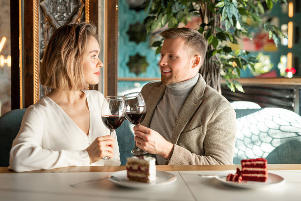Handsome Caucasian man and beautiful woman sitting together at cafe table clinking glasses with red wine looking at each other - Photo, Image