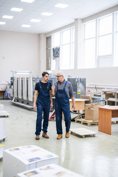 Старшие и молодые работники типографии в синем комбинезоне пересекают современную мастерскую и обсуждают работу
 - Фото, изображение
