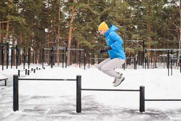 Widok z boku młodzieńca w niebieskiej kurtce trening przy użyciu poziomych prętów jako przeszkody do skakania w zimie - Zdjęcie, obraz