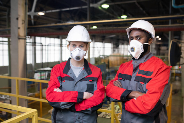 Portrait de travailleurs sérieux portant des masques respiratoires et des casques de sécurité debout avec des bras croisés sur un pont-charpente dans un atelier d'usine
 - Photo, image