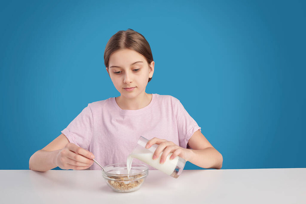 Adolescente sentada à mesa e adicionando leite em cereais enquanto toma café da manhã, fundo azul
 - Foto, Imagem