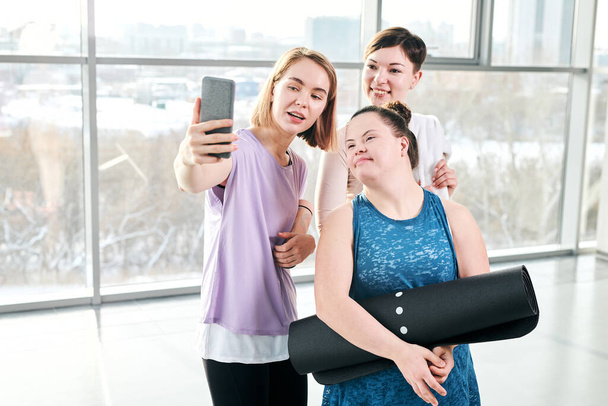 Joven instructor de fitness feliz y dos mujeres activas mirando la cámara del teléfono inteligente mientras se hace selfie después del entrenamiento
 - Foto, Imagen