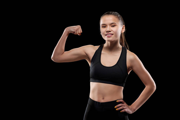 Glückliche junge Sportlerin in schwarzer Aktivkleidung, die dich ansieht und dabei ihre körperliche Stärke und Muskeln zeigt - Foto, Bild