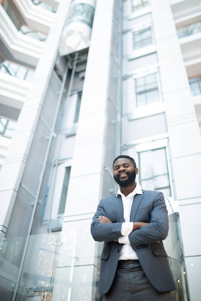 Πορτρέτο του χαρούμενου επιτυχημένου νεαρού Αφροαμερικανού επιχειρηματία με κοστούμι στέκεται με σταυρωμένα χέρια στο λόμπι του γραφείου - Φωτογραφία, εικόνα