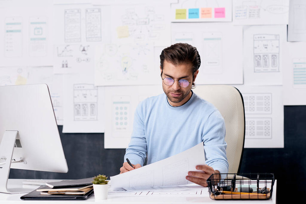 Горизонтальный снимок молодого кавказца в очках, сидящего за столом офиса и работающего с бумагами
 - Фото, изображение