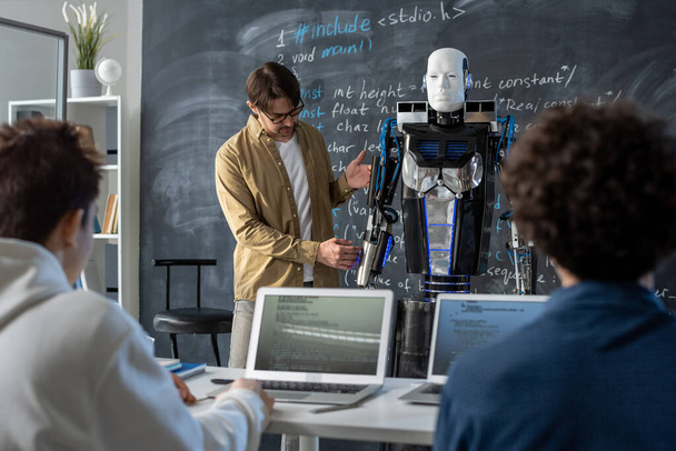 Dozent der Technischen Universität steht neben Automatisierungsroboter an Tafel, während er Innovationen einer Gruppe von Studenten im Seminar vorstellt - Foto, Bild