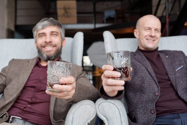Счастливые успешные мужчины в модных нарядах, сидящие в креслах и пьющие алкоголь вместе
 - Фото, изображение