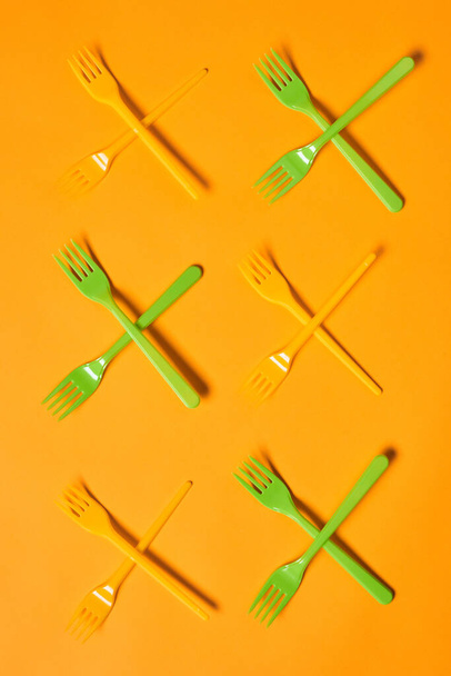Κάθετη εννοιολογική επίπεδη lay shot από X σήματα κατασκευασμένα από ανοιχτό πράσινο φως μιας χρήσης και πορτοκαλί πλαστικά πιρούνια σε πορτοκαλί φόντο - Φωτογραφία, εικόνα