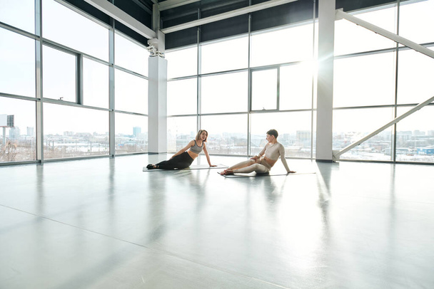 Dos jóvenes deportistas en ropa deportiva sentadas en alfombras después del entrenamiento y hablando contra grandes ventanales en el centro de ocio
 - Foto, Imagen