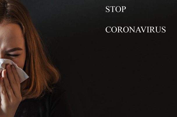 Eine kranke junge Frau mit einer runzeligen Nase, die ihre Nase pustet und runzelt, hat sich erkältet, niest in Gewebe, hustet und hat Grippe. Auf schwarzem Hintergrund die Aufschrift "Stop Coronavirus"". - Foto, Bild