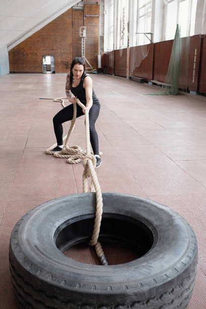 Young Caucasian female athlete pulling heavy tire with battle rope while having cross training workout in sports hall - Photo, Image