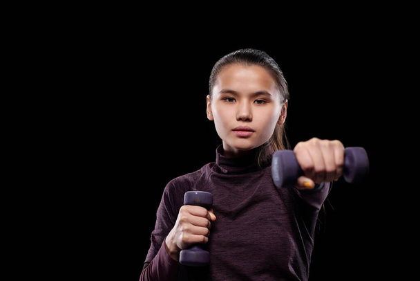 Aktivní sportovní dívka s činky natahování paže dopředu před kamerou při cvičení na černém pozadí - Fotografie, Obrázek