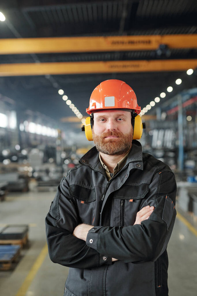 Feliz jovem trabalhador industrial barbudo em fones de ouvido de proteção e capacete em pé na frente da câmera na fábrica
 - Foto, Imagem