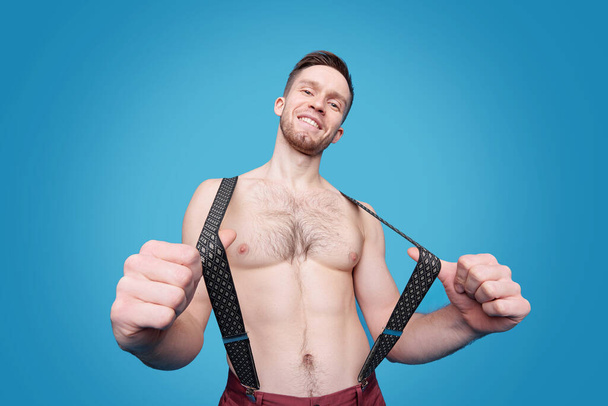 Portret van vrolijke sexy man met haar op borst trekken bretels tegen blauwe achtergrond - Foto, afbeelding