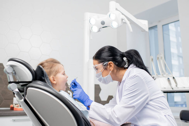 Joven dentista morena en blanco, guantes, anteojos y máscara con gancho dental y espejo durante el examen oral
 - Foto, imagen