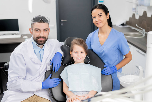 Feliz niña en sillón con su dentista y su asistente de pie cerca mirándote en la oficina de odontología
 - Foto, imagen