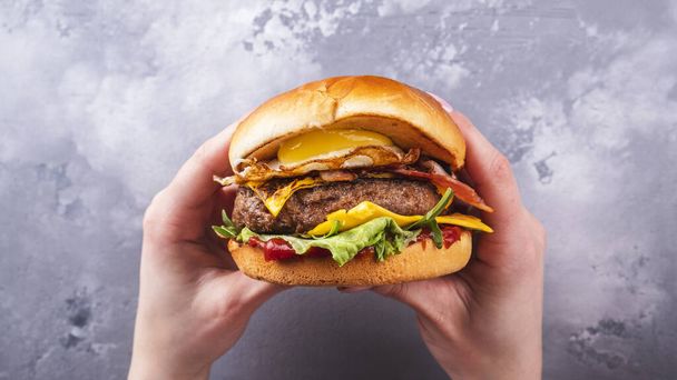 Weibliche Hände halten leckere Rindfleisch-Burger mit Spiegelei und Speck in der Hand. Ansicht von oben. Amerikanische Lebensmittel - Foto, Bild