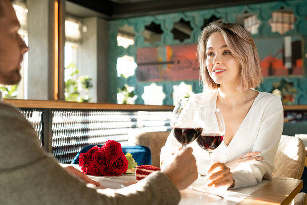 Horizontal over-the-shoulder shot of young adult man and woman in love sitting in restaurant clinking glasses with red wine - Photo, Image