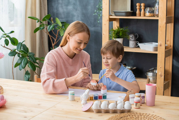 Curious boy sitting at wooden table and painting eggs with gouache while spending time with mother - Photo, Image