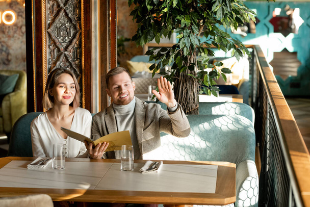 Horizontal shot of beautiful couple sitting in modern restaurant with menu book calling waiter - Photo, Image