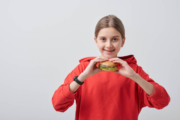 Retrato de adolescente sorridente em camisola vermelha comendo hambúrguer contra fundo branco
 - Foto, Imagem