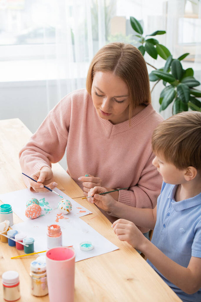 Mladá matka sedí u stolu a malovat vejce se synem, zatímco oni dělají dekorace na velikonoční oslavu - Fotografie, Obrázek