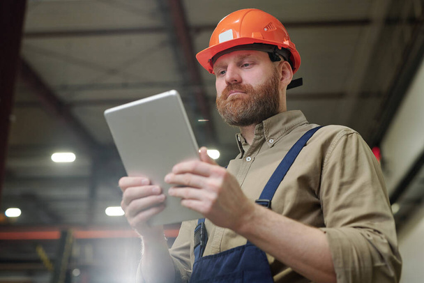 Νεαρός μηχανικός με κόκκινη γενειάδα κρατώντας tablet μπροστά από τον εαυτό του, ενώ κοιτάζοντας μέσα από online δεδομένα κατά τη διάρκεια της εργασίας - Φωτογραφία, εικόνα