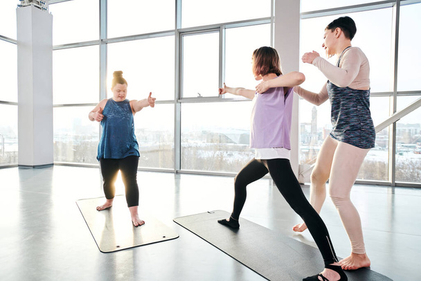 Treinador de ioga jovem ajudando duas mulheres ativas a se exercitar em esteiras, enquanto em pé por um deles no centro de lazer
 - Foto, Imagem